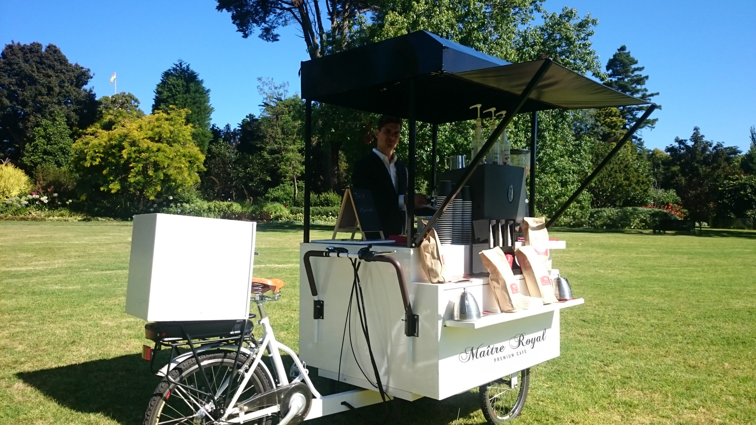 Unser weißes Kaffee-Fahrrad bei einer Veranstaltung, im Park während einer öffentlichen Veranstaltung. Sommerliches Flair. Einer unserer Baristas steht lächelnd hinter dem Coffee-Bike. Unvergleichlicher Geschmack für Ihre Veranstaltung.