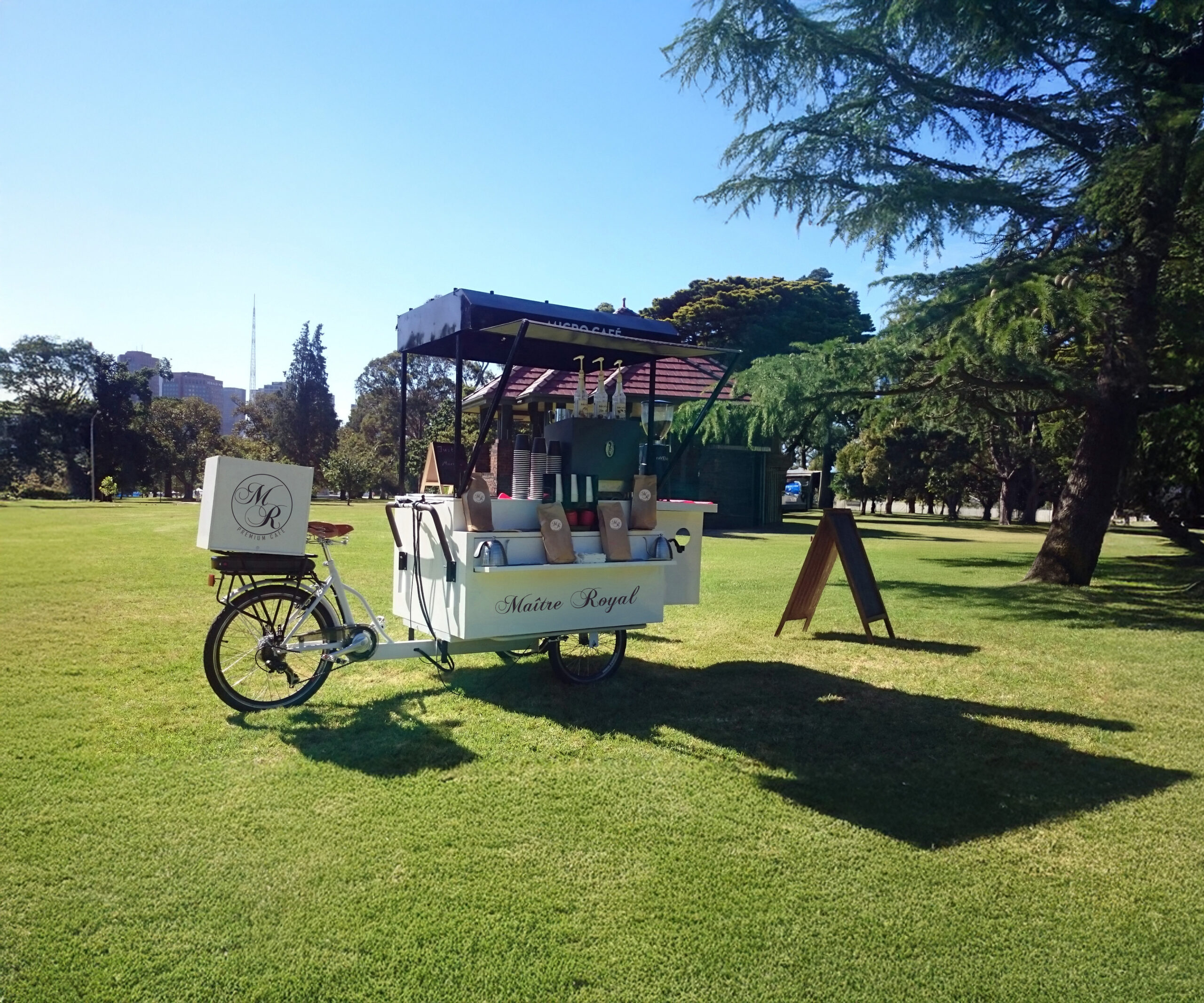 Unser weißes Kaffee-Fahrrad bei einer Veranstaltung, im Park während einer öffentlichen Veranstaltung. Sommerliches Flair. Einer unserer Baristas steht lächelnd neben dem Coffee-Bike. Unvergleichlicher Geschmack für Ihre Veranstaltung.