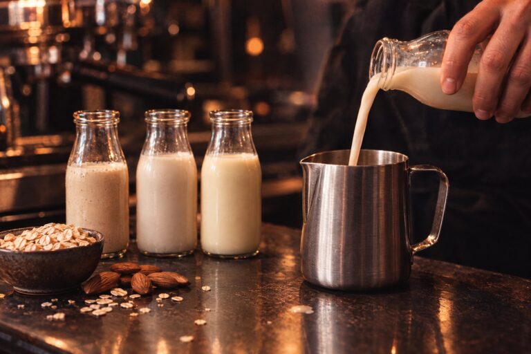 Drei Flaschen mit Pflanzendrinks, Haferflocken und Mandeln, ein Barista gießt Haferdrink in die Milchkanne