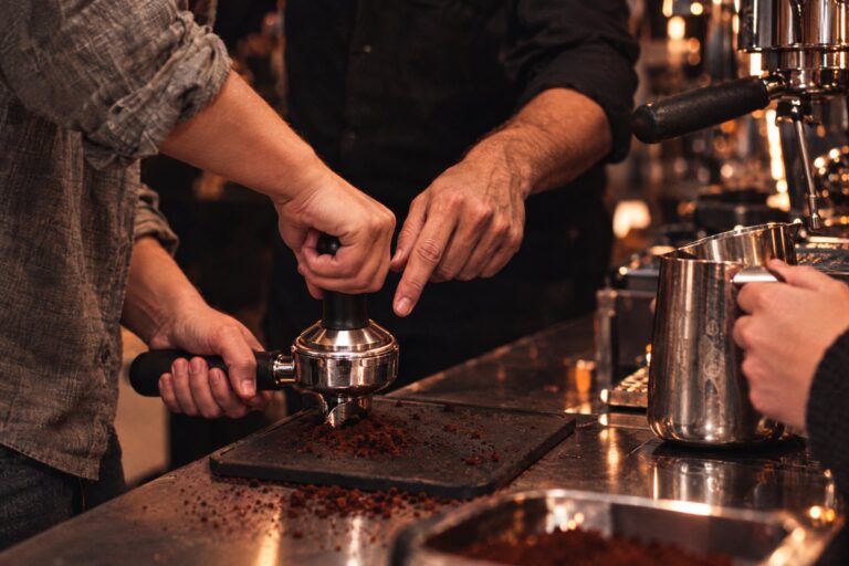 Teilnehmer eines Barista-Trainings tampen Kaffee im Siebträger, ein Barista korrigiert die Haltung