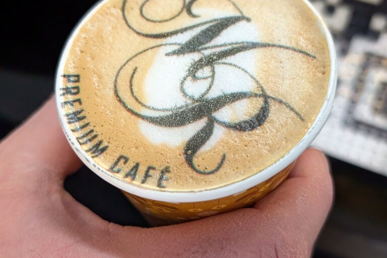 Hand hält einen Kaffeebecher, auf dessen Milchschaum das Logo von Maitre Royal gedruckt ist