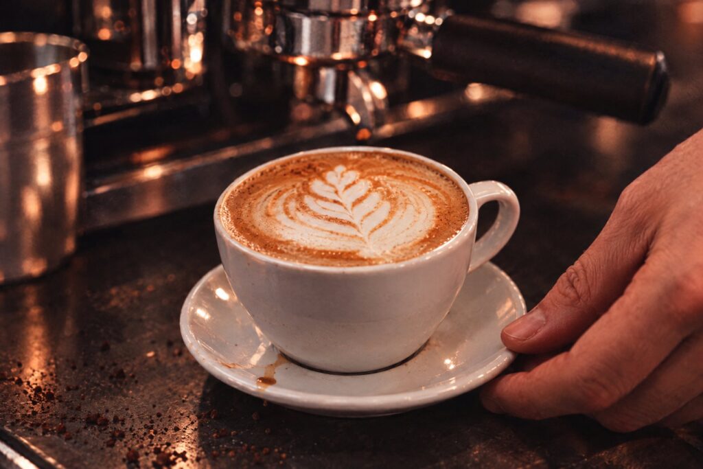 Cappuccino mit gegossener Latte-Art-Rosette auf Untertasse, ein Barista stellt ihn auf die Theke