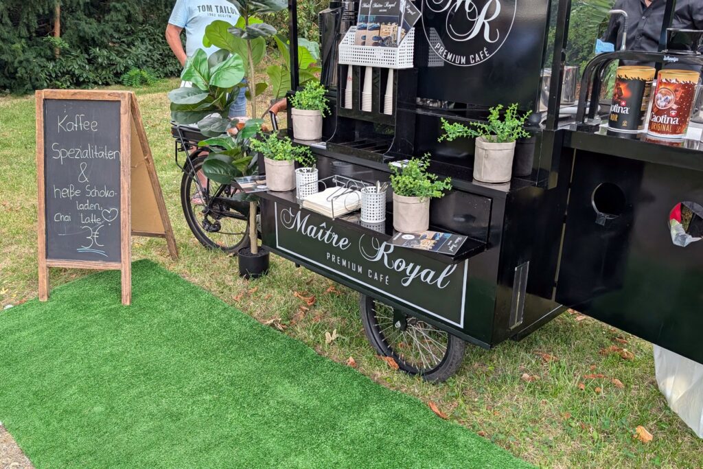 Kaffeerad von Maitre Royal auf Kunstrasen im Schlossgarten, daneben eine Kreidetafel mit der Getränkekarte
