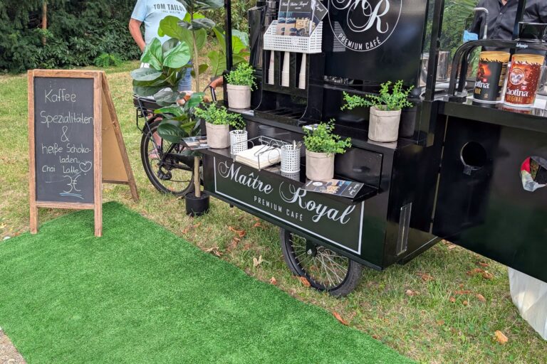 Kaffeerad von Maitre Royal auf Kunstrasen im Schlossgarten, daneben eine Kreidetafel mit der Getränkekarte