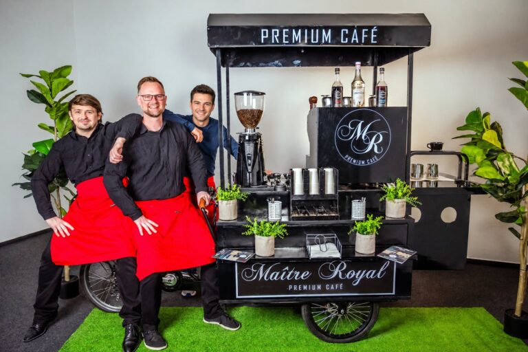 Drei Baristas von Maitre Royal in schwarzen Hemden und roten Schürzen vor dem schwarzen Kaffeerad mit Premium-Café-Dach