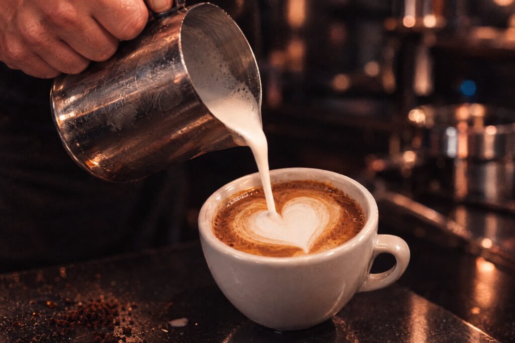 Barista gießt aufgeschäumte Milch aus einer Edelstahlkanne in einen Cappuccino, ein Herz entsteht