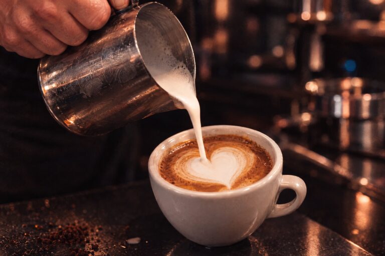 Barista gießt aufgeschäumte Milch aus einer Edelstahlkanne in einen Cappuccino, ein Herz entsteht
